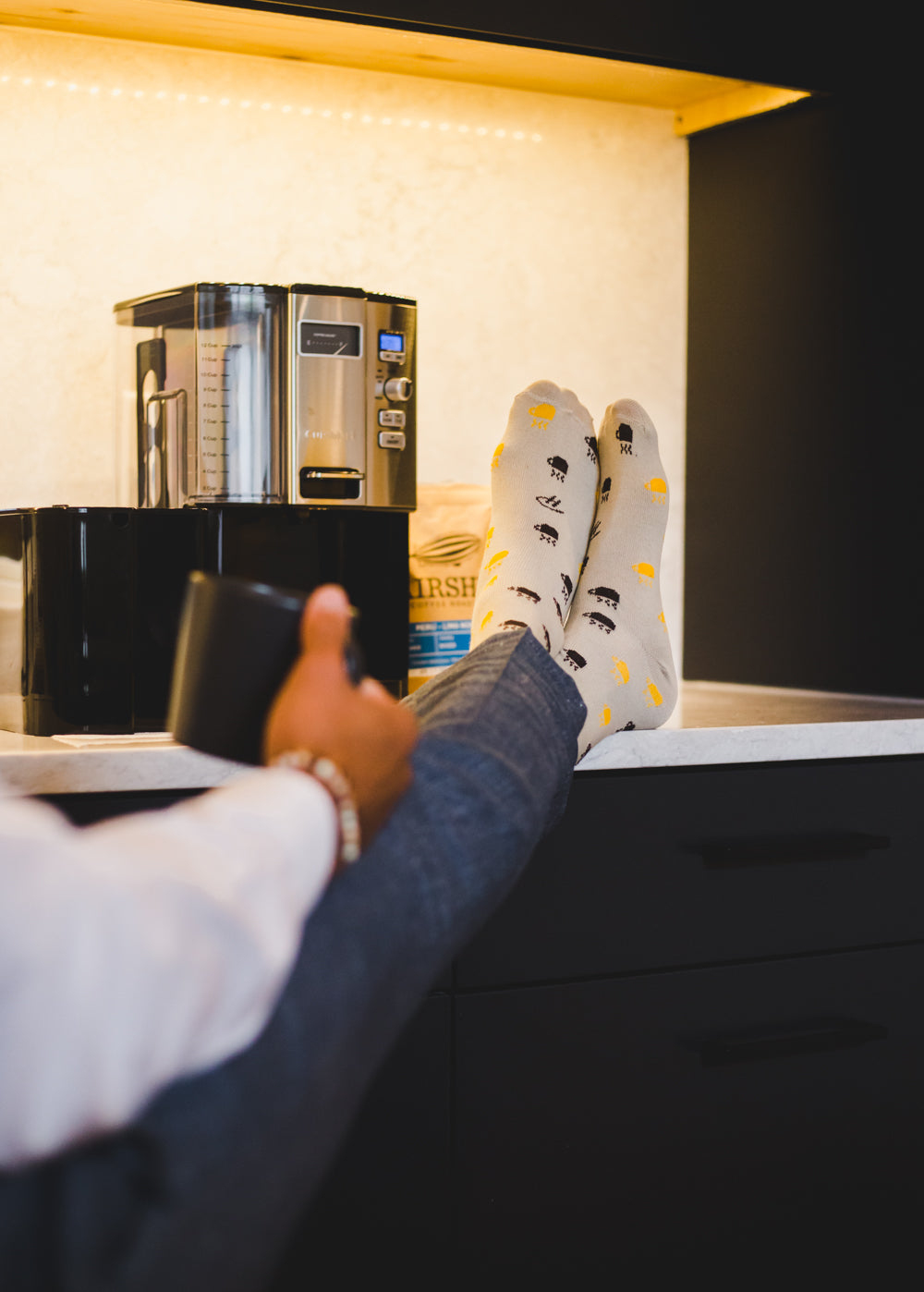 light grey over the calf dress socks with colored hot mug print, a black coffee mug, a coffee machine, blue jeans