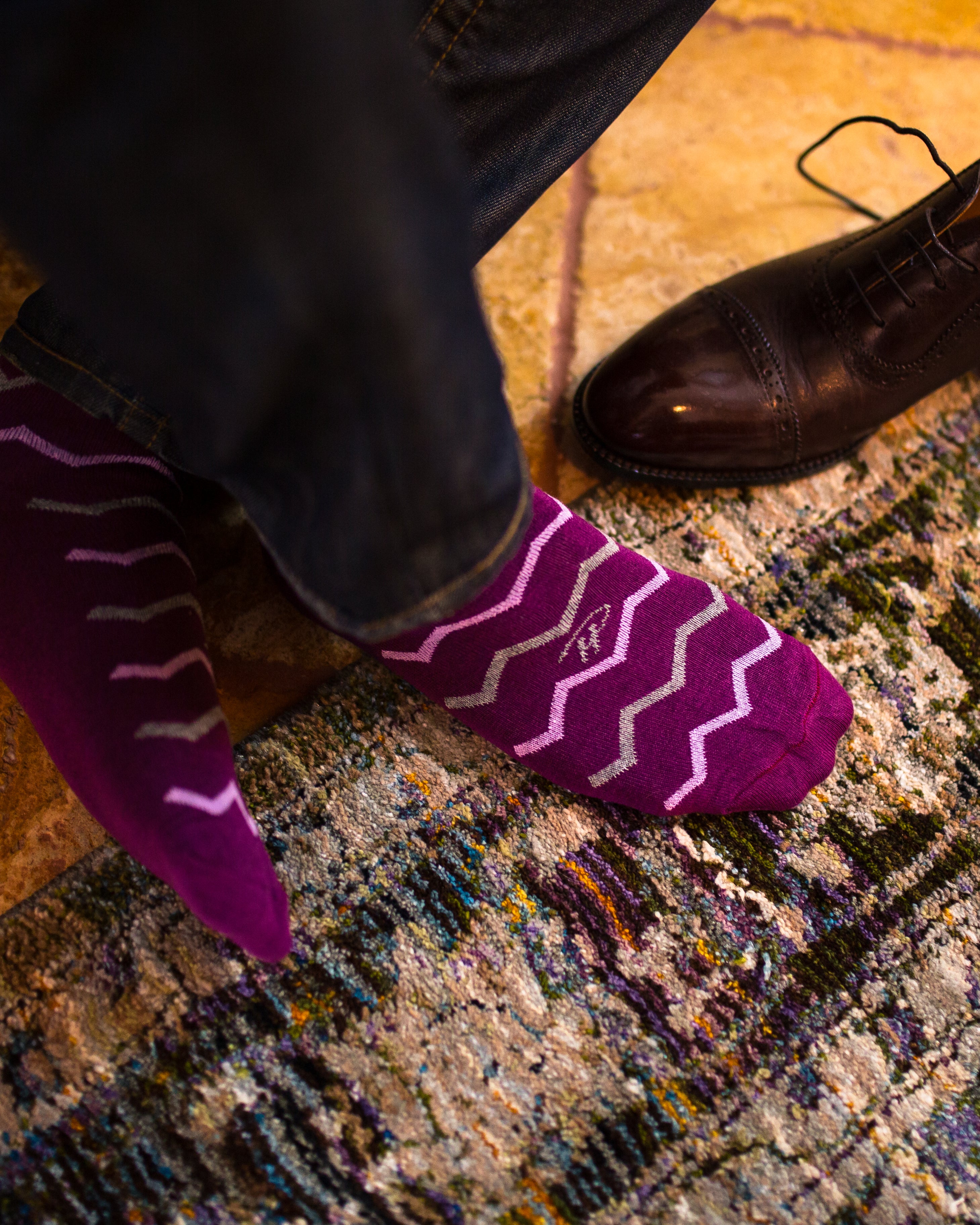 purple over the calf dress socks with grey and pink zigzag lines, brown dress shoe on the floor, blue jeans